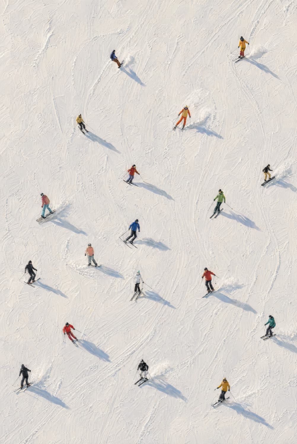 Aerial view of around twenty skiers and snowboarders descending a fresh-powder slope; figures wear warm vintage-toned jackets — red, yellow, orange, mustard, green — casting long soft shadows across the matte-textured snow.