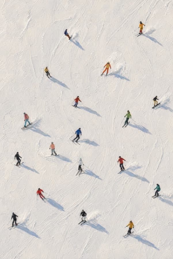 Aerial view of around twenty skiers and snowboarders descending a fresh-powder slope; figures wear warm vintage-toned jackets — red, yellow, orange, mustard, green — casting long soft shadows across the matte-textured snow.